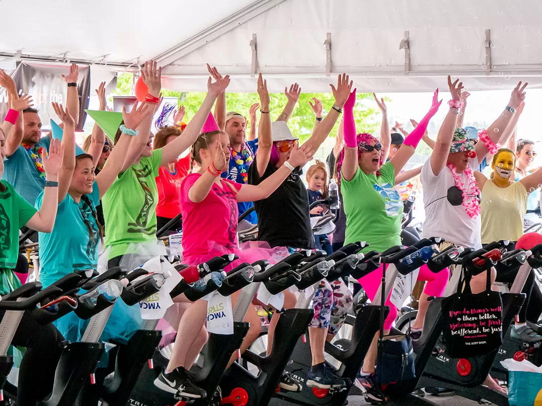 A group of 30 people are dressed up in costumes and bright colors, and sitting on stationary bikes in an outdoor class. They are all smiling and cheering and holding their hands up in the air. 