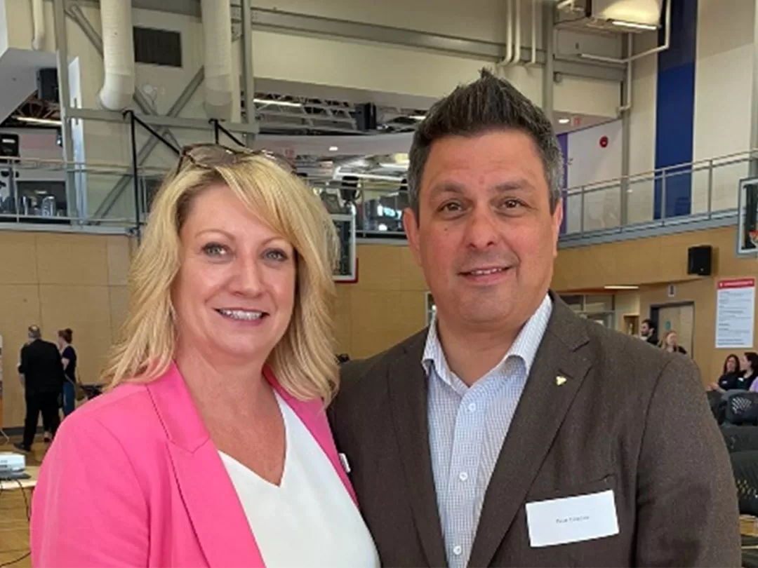 A professional woman and man are standing together smiling at the camera with an event taking place behind them in a gymnasium. 