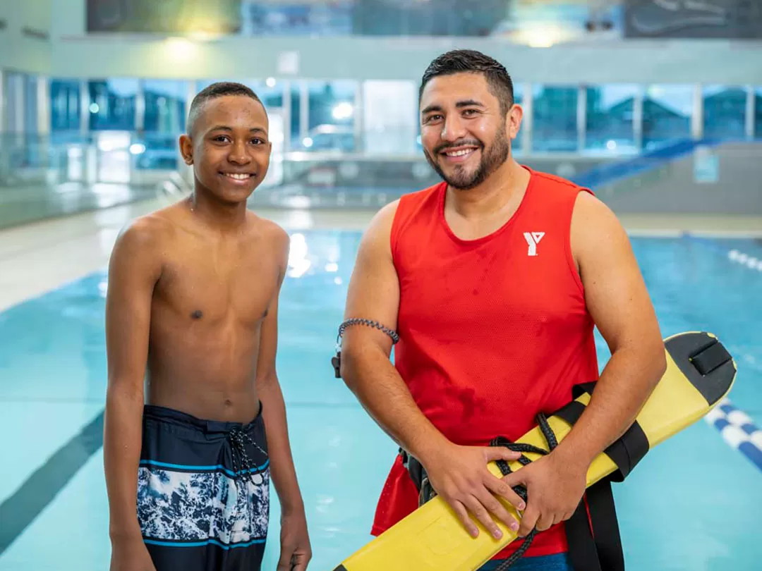 A young teen in swim trucks stands beside a male lifeguard with dark hair in front of a pool. They are smiling at the camera. 