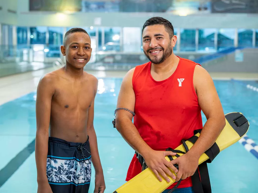 A young teen in swim trucks stands beside a male lifeguard with dark hair in front of a pool. They are smiling at the camera.