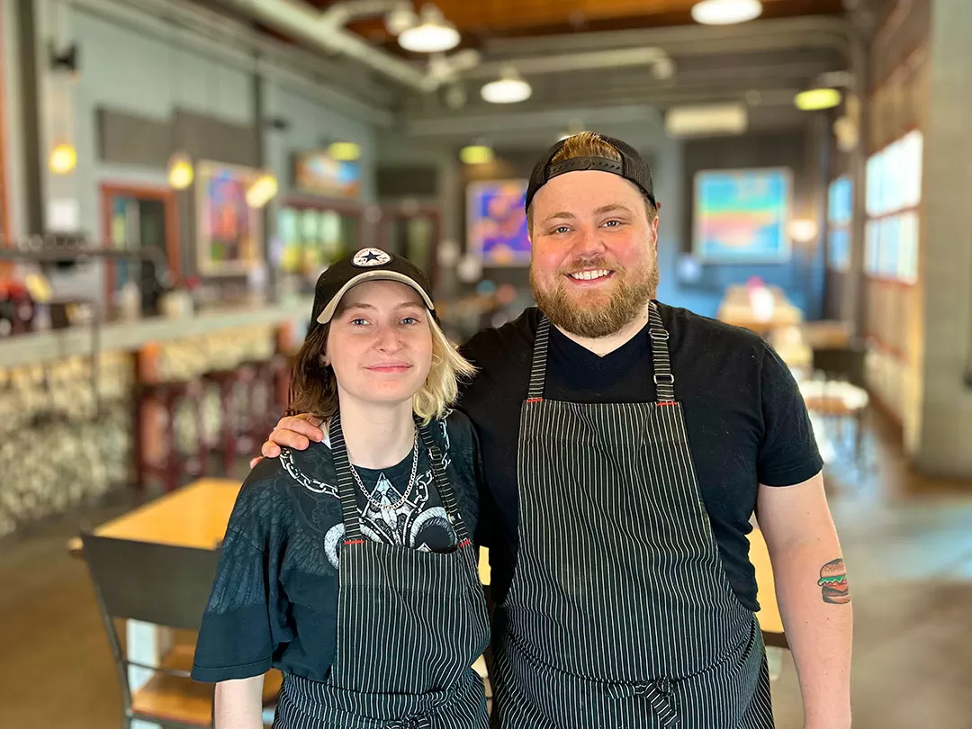 A young man and woman in black striped chef aprons are standing in a restaurant with their arms around one another and smiling at the camera. 