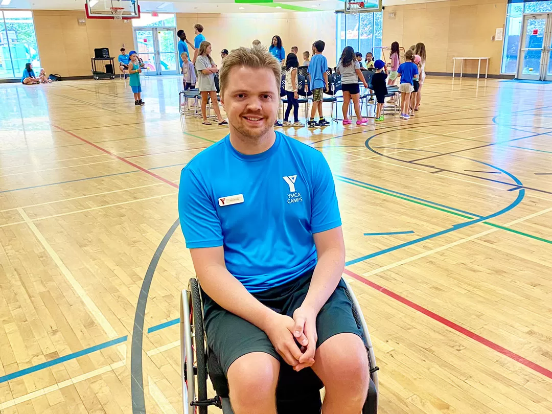 A young blonde man in a blue shirt and a wheelchair is smiling at the camera in a gymnasium. There's a group of 20 kids and leaders playing a game in the background.