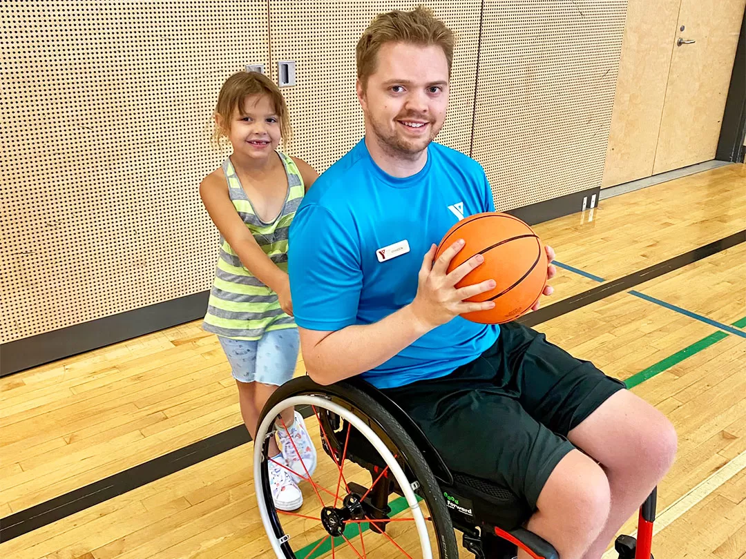 A young blonde man in a blue shirt and a wheelchair is smiling at the camera and holding a basketball in a gymnasium. There's an 8 year old girl holding the back handles of the chair also smiling at the camera. 