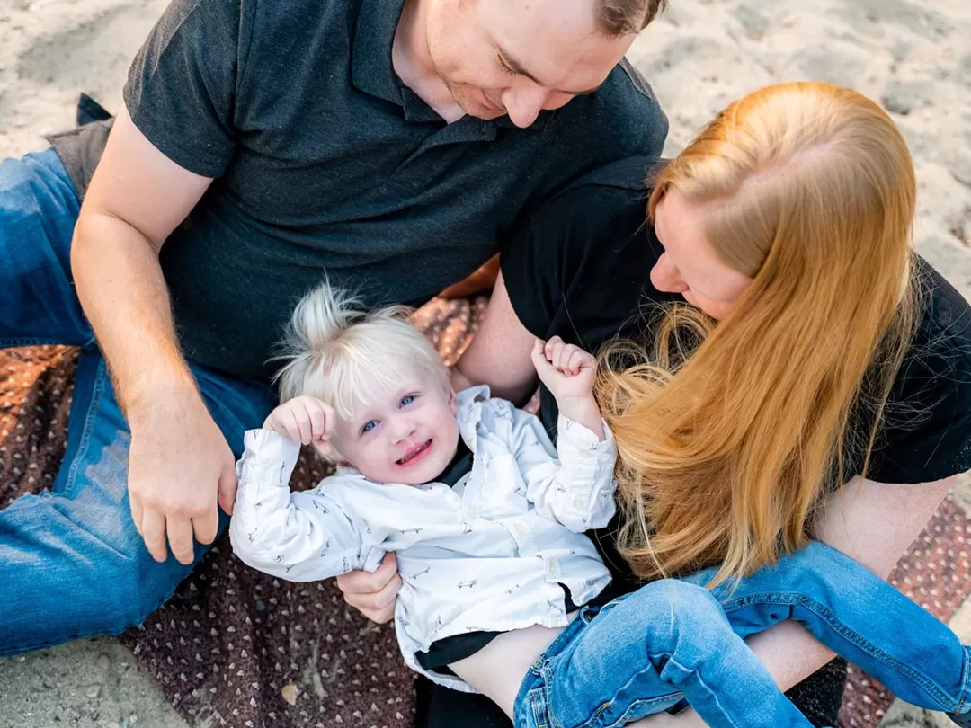 A mother and folder are holding their 2 year old boy and looking down at him as they sit on a beach. The little boy has blue eyes, blonde hair, and is smiling up at the camera.