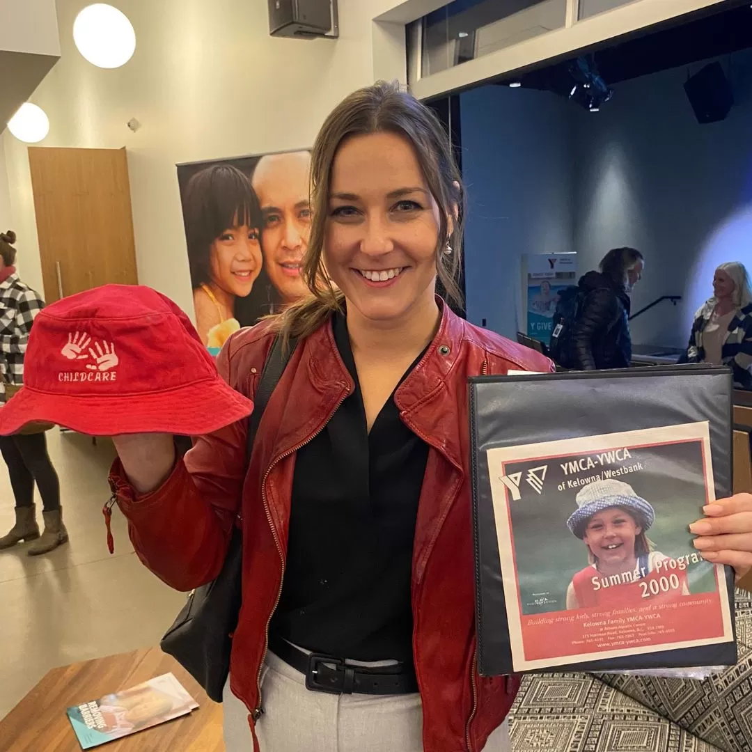 A young professional woman with dark hair is smiling at the camera and holding an older YMCA child’s bucket hat in one hand, and a photo of herself as a child in an old YMCA program advertisement in the other.