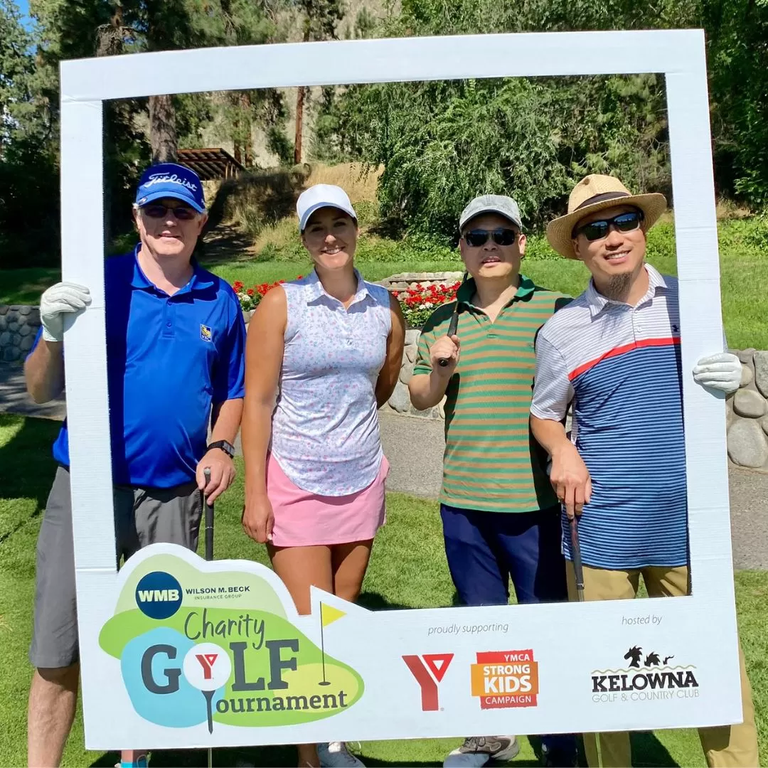 A group of  golfers (1 female and 1 male) are standing outside on a golf course and smiling at the camera and holding a large white photo frame around them.