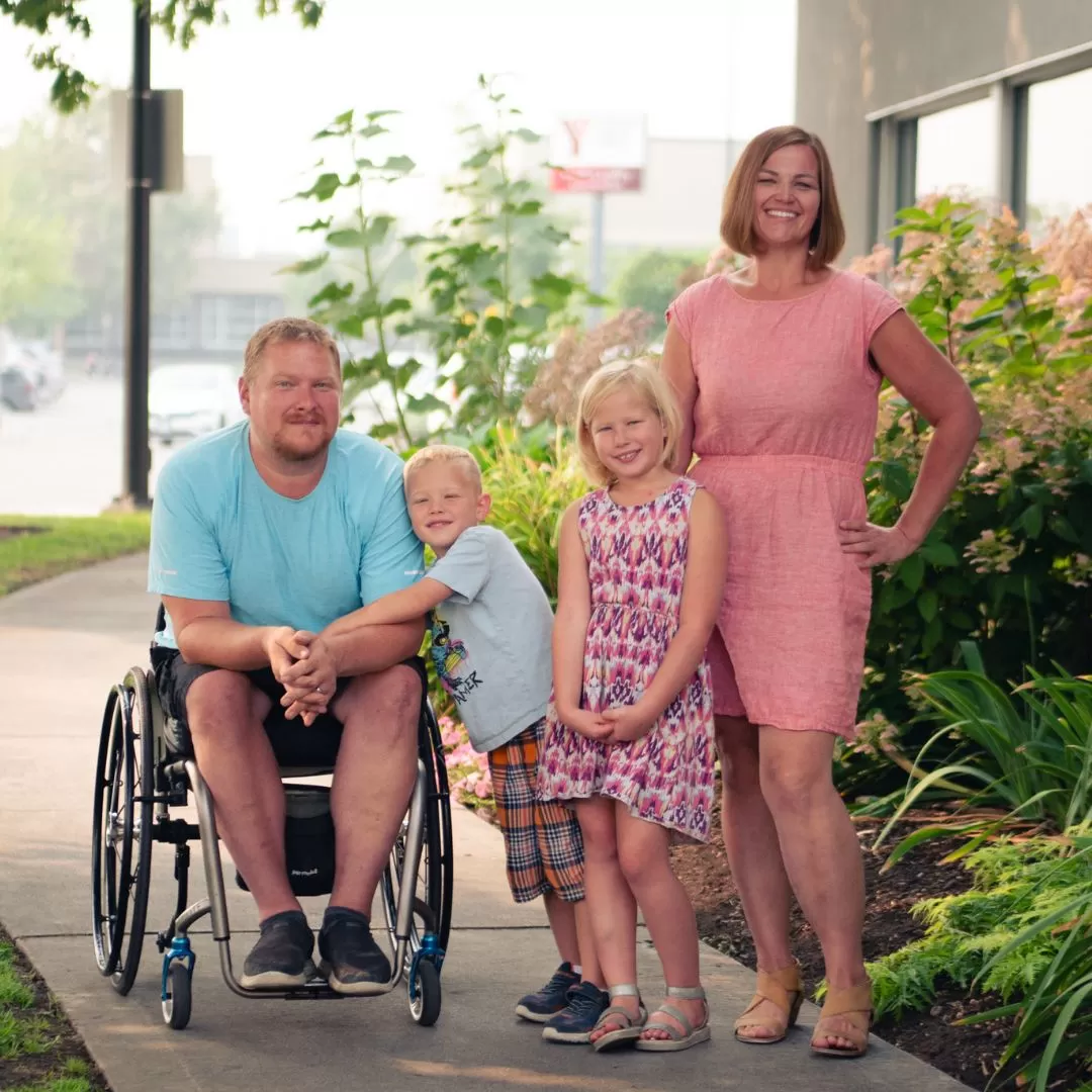 A family of 4 with two young children under the age of 7 are standing together outside in the sunshine and smiling at the camera.