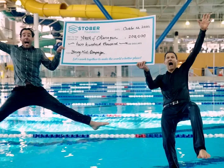 2 men jumping in a pool with a huge cheque