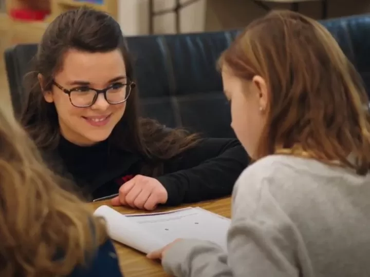 A young woman with glasses, brown hair and brown eyes is kneeling in front of a classroom table smiling at a 7 year old girl sitting at the table. The young girl has shoulder length brown hair, and her back is to the camera as she looks down at an open notebook in front of her.  