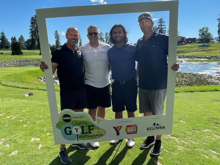  Four men in golf attire are standing outside on a golf course on a beautiful sunny day. They are all smiling at the camera and surrounded by a white frame they are holding around themselves. The men range in age from 30-55.