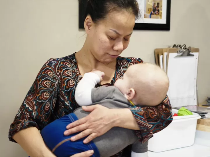 A woman in a patterned blouse is holding baby boy to her chest at a child care centre. The baby is asleep with his hand on her neck.