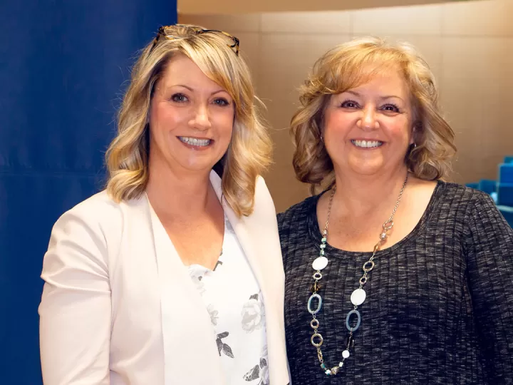 Two women are standing side by side and smiling at the camera. Allyson Graf is on the left with mid length blonde hair and a cream jacket, Sharon Peterson is on the right with shoulder length auburn hair and a grey sweater. 
