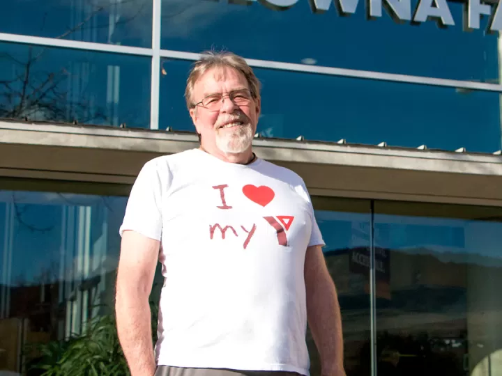 A man with glasses and a grey goatee is standing in front of the Kelowna Family Y and smiling at the camera. He’s wearing a white shirt that says ‘I love my Y’.