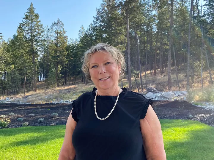 A woman with short blonde hair, wearing a black dress and pearls is smiling at the camera. There is a forest behind her and the sun is shining. 