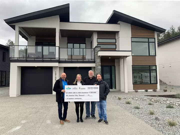 There are four adults standing in the driveway in front of a brand new modern luxury home. They are all smiling at the camera and holding a donation cheque of $150,000 to the KGH Foundation and YMCA of Southern Interior BC.