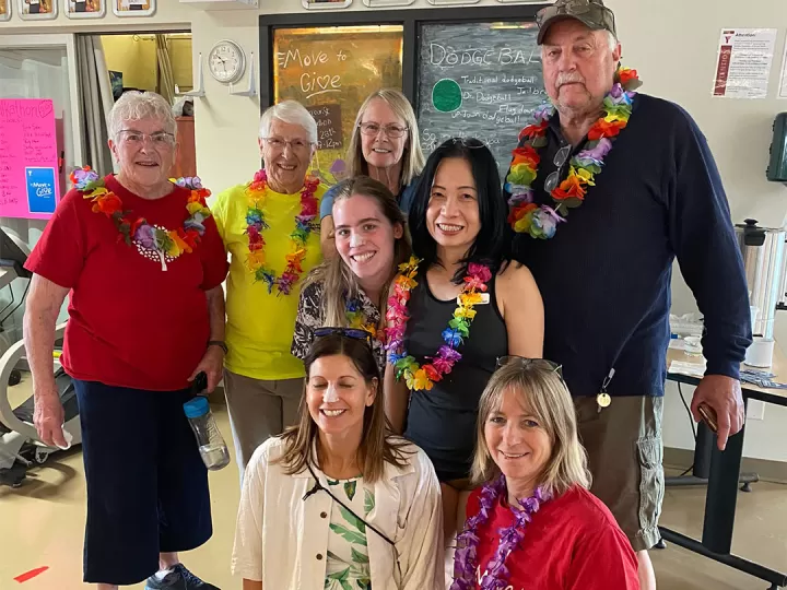 A group of 8 seniors and adults are standing together in a gym smiling at the camera. They are all wearing athletic clothes and ‘tourist wear’ including Hawaiian lays, tropical shirts and bright colors. 