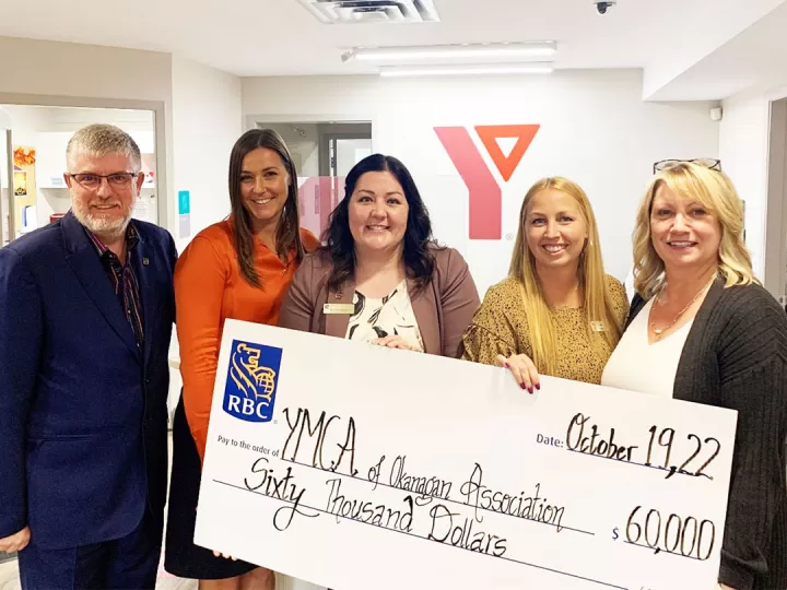 5 professionals of various ages in business clothing are standing in a YMCA office, holding a large donation cheque and smiling at the camera. 