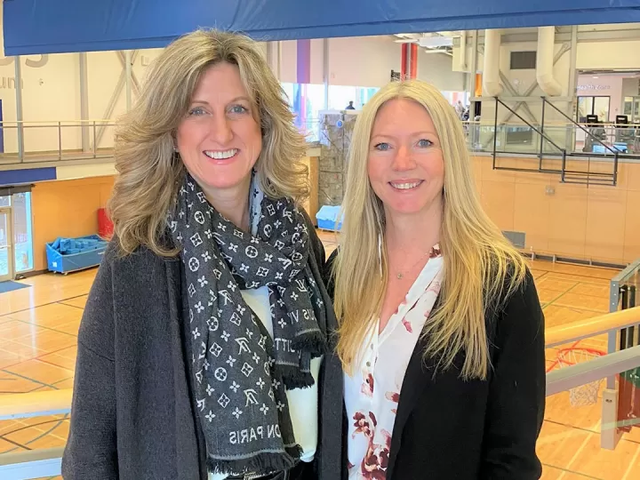 Two blonde women in professional clothing are standing in front of a gymnasium and smiling at the camera. 