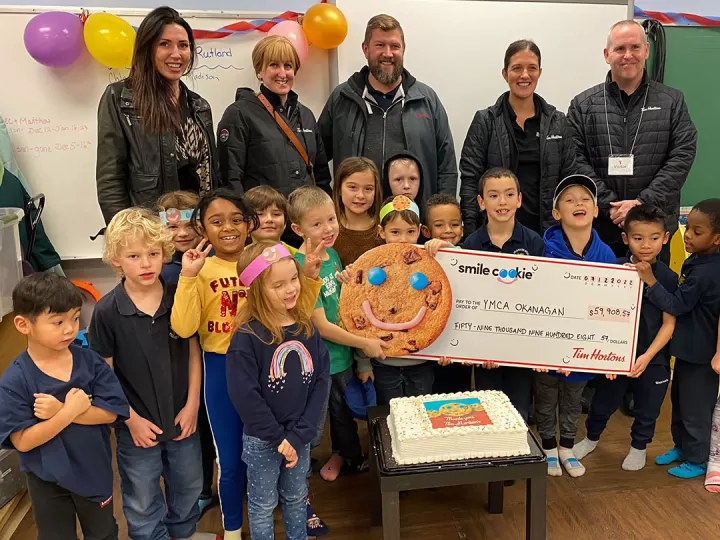 Five adults are standing in a classroom smiling at the camera. In front of them a group of 15 children aged 5-7 are smiling and holding a large donation cheque made out to the YMCA.