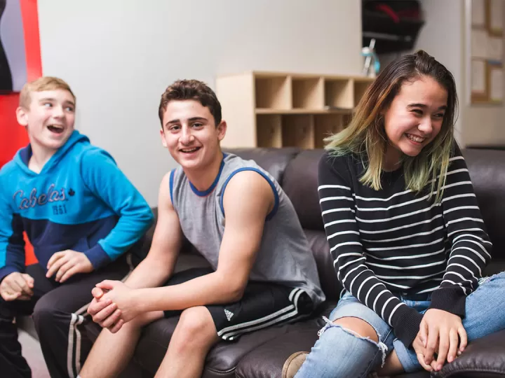 Three teens are sitting on a couch, smiling in different directions as they interact with people off camera. Two on the left are boys and there is one girl on the right.