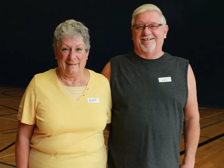 An older woman and man are standing in a gymnasium with their arms around one another smiling at the camera. The woman has grey hair and a yellow t-shirt while the man has white hair, glasses, and a grey sleeveless workout shirt on. 