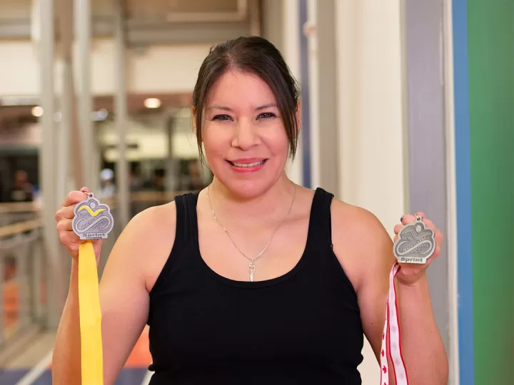 A woman with dark hair pulled back in workout clothes is holding two medals up and smiling at the camera. There is a gymnasium in the background. 