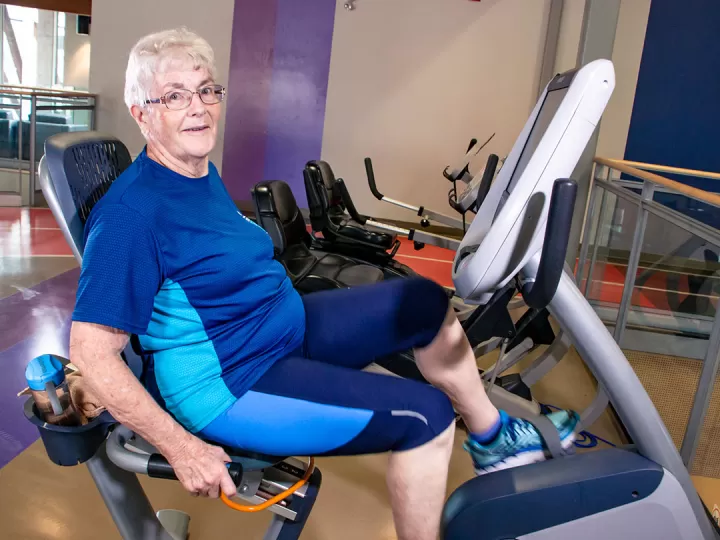 A woman with glasses, short white hair, and blue workout clothes is sitting on a laid back cycle machine in a gym and smiling at the camera. 