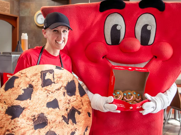 A YMCA mascot is holding an open box of smile cookies and posing beside a young female Tim Hortons Store Owner who is dressed as a large cookie. 
