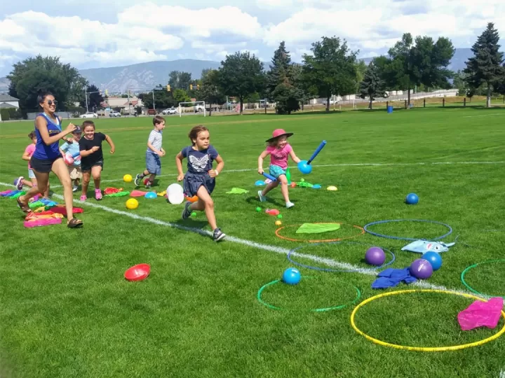 A female YMCA camp leader runs through an outdoor obstacle course with 6 children, aged 7-9, outside on a sunny day. 