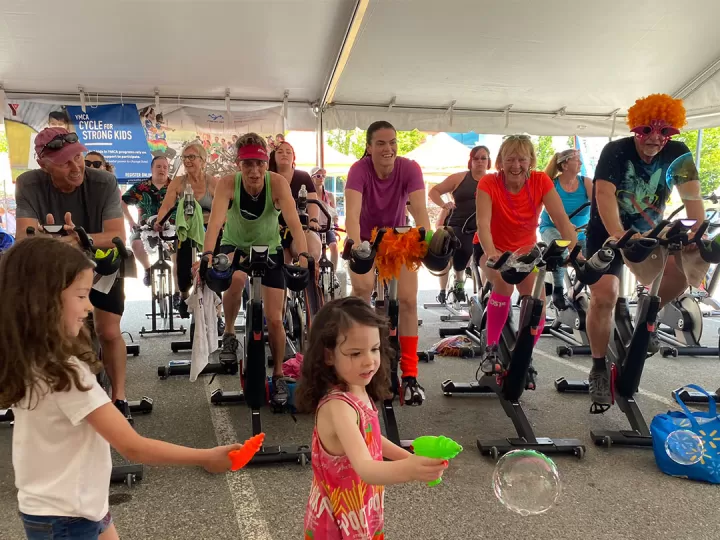 Two young girls with brown hair are playing with water guns and bubbles. Behind them a group of 14 adults dressed in costumes and athletic wear are riding cycle bikes in an outdoor tent and smiling. 