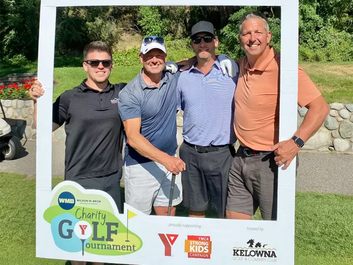 Four men in golf attire are holding clubs and smiling at the camera on a golf course.