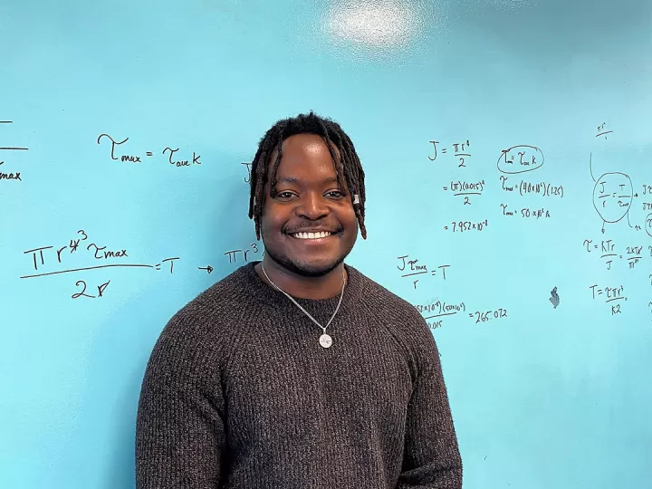 A young man with short black thin dreadlocks in a brown sweater is smiling at the camera in front of a teal board with mathematical equations scribbled on it.