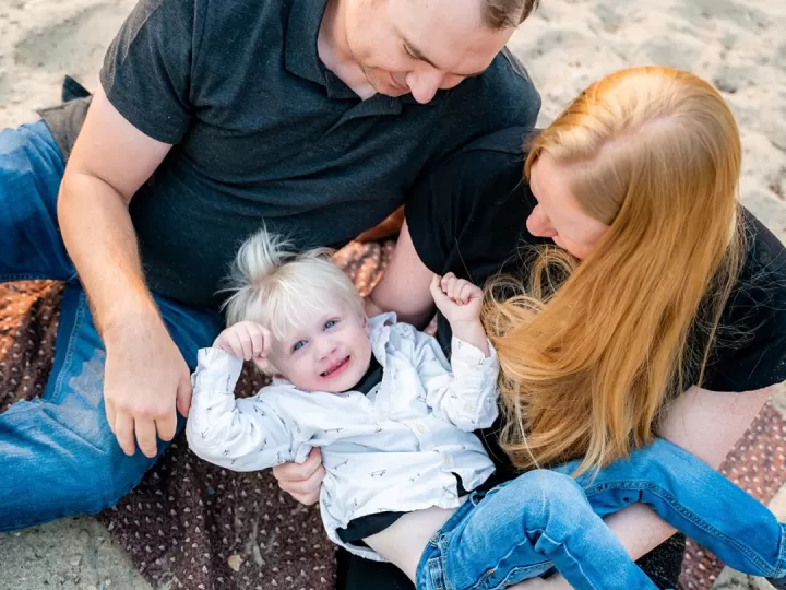 A mother and folder are holding their 2 year old boy and looking down at him as they sit on a beach. The little boy has blue eyes, blonde hair, and is smiling up at the camera.