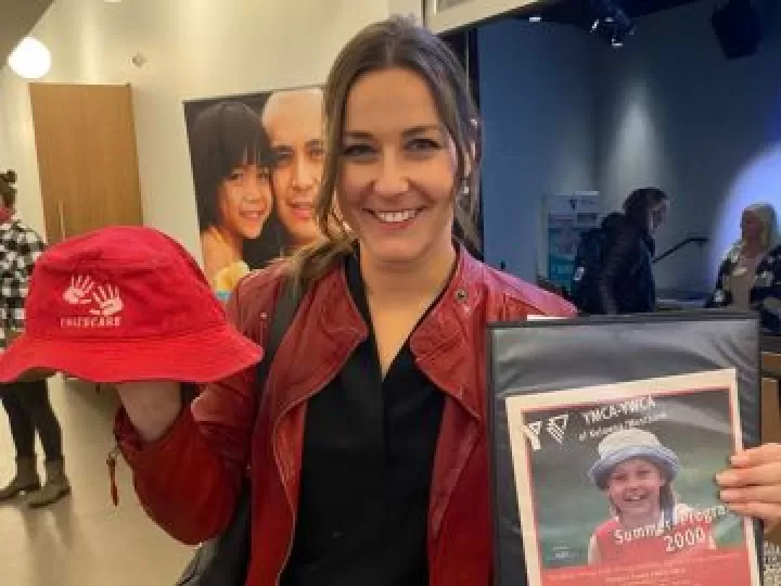 A young professional woman with dark hair is smiling at the camera and holding an older YMCA child’s bucket hat in one hand, and a photo of herself as a child in an old YMCA program advertisement in the other.