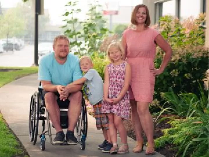 A family of 4 with two young children under the age of 7 are standing together outside in the sunshine and smiling at the camera.