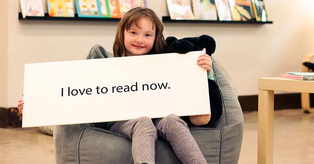 7 year old girl sitting in a grey bean bag chair with a black teddy bear smiling. She is holding a large white sign that says 'I love to read now' and there is a library or classroom with books in the background. 