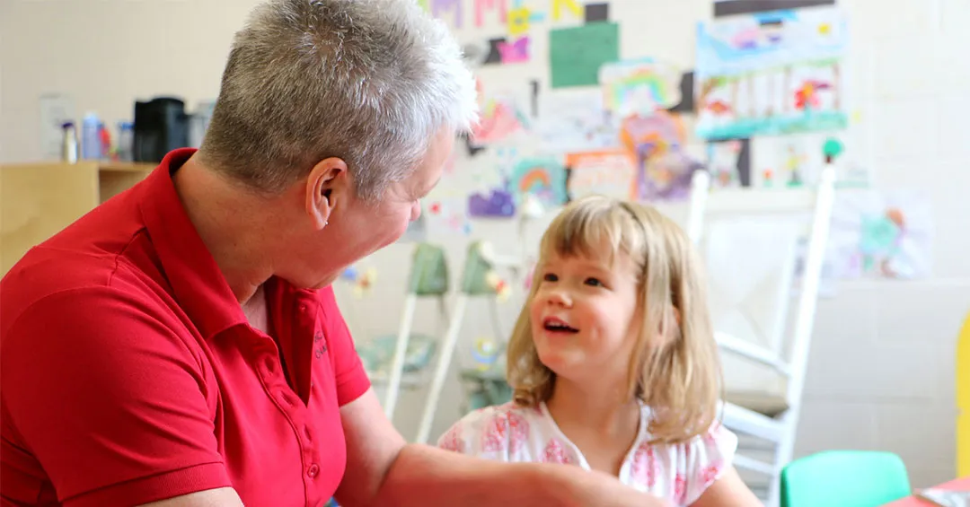 Woman in red shirt and 4 year old girl sitting at a table with crafts looking at oneanother and smiling