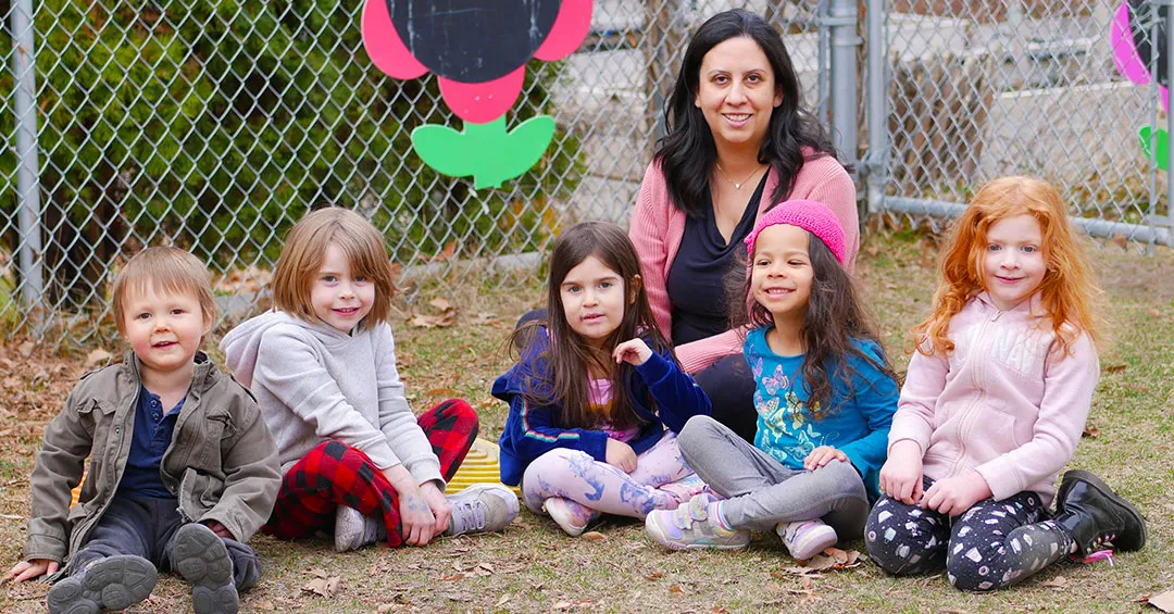 A woman is sitting in a playground with 5 children between 3-5 sitting on the ground in front of her. Everyone is smiling at the camera.