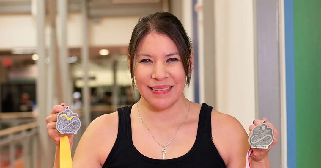 A woman in a black tank with dark hair is holding up two triathlon medals and smiling at the camera. She's standing in front of a gymnasium. 