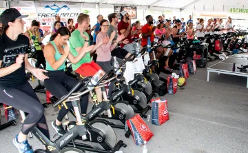 Large outdoor Cycle Class taking place under a tent with roughly 50 brightly dressed cyclists smiling and cheering