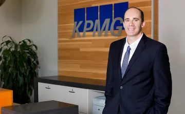 A professional shot of a man in a suite with light blue tie standing with his hands in his pockets smiling at the camera. There is a receptionist desk behind him with a sign that reads KPMG.