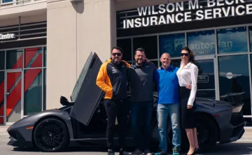 Three men and one woman are standing in front of a high-end grey sports car with suicide doors open. They have their arms around one another and are smiling at the camera. The photo is taken outside on a sunny day with offices behind them. 