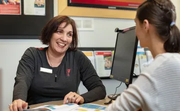 Middle aged woman with chin length dark hair in a grey YMCA shirt is sitting at a customer service desk in front of a computer and smiling at young female customer sitting in front of her.  