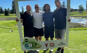  Four men in golf attire are standing outside on a golf course on a beautiful sunny day. They are all smiling at the camera and surrounded by a white frame they are holding around themselves. The men range in age from 30-55.