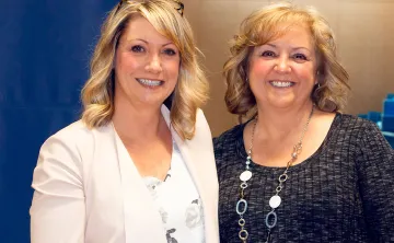 Two women are standing side by side and smiling at the camera. Allyson Graf is on the left with mid length blonde hair and a cream jacket, Sharon Peterson is on the right with shoulder length auburn hair and a grey sweater. 