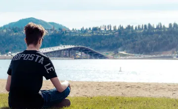A young man with short dark hair, jeans, sun glasses and a black t shirt is sitting cross legged looking out at a beach. The photo is taken behind him and he is looking out at the water, with a bridge full of cars crossing the lake in the distance. 
