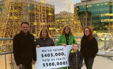 Four adults and one child are standing outside in front of an enormous Christmas tree made of Christmas lights. They are all smiling at the camera and holding a sign that reads 'We've raised $403,00, help us reach $500,000!'. 