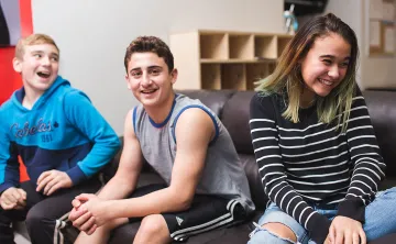 Three teens are sitting on a couch, smiling in different directions as they interact with people off camera. Two on the left are boys and there is one girl on the right.