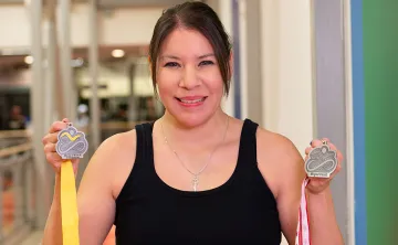 A woman with dark hair pulled back in workout clothes is holding two medals up and smiling at the camera. There is a gymnasium in the background. 