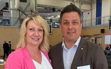 A professional woman and man are standing together smiling at the camera with an event taking place behind them in a gymnasium. 